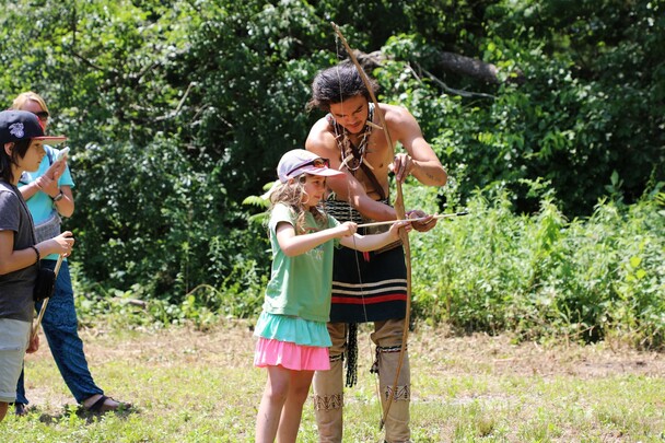 An Indigenous Educator helps a young girl learn how to shoot an arrow on the Historic Patuxet Homesite.