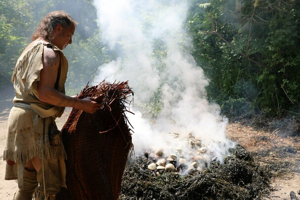 Indigenous Educator dressed in regalia covers a traditional clambake with water soaked mats.