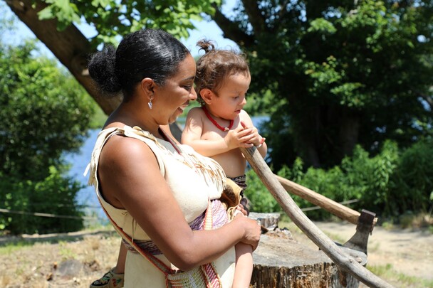 A Museum Educator dressed in Wampanoag regalia holds a toddler boy in regalia has he tries to scrape the mishoon on the Homesite.