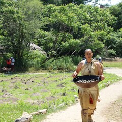 Museum Educator carries a wooden bowl filled with clams through the planting field on the Historic Patuxet Homesite.
